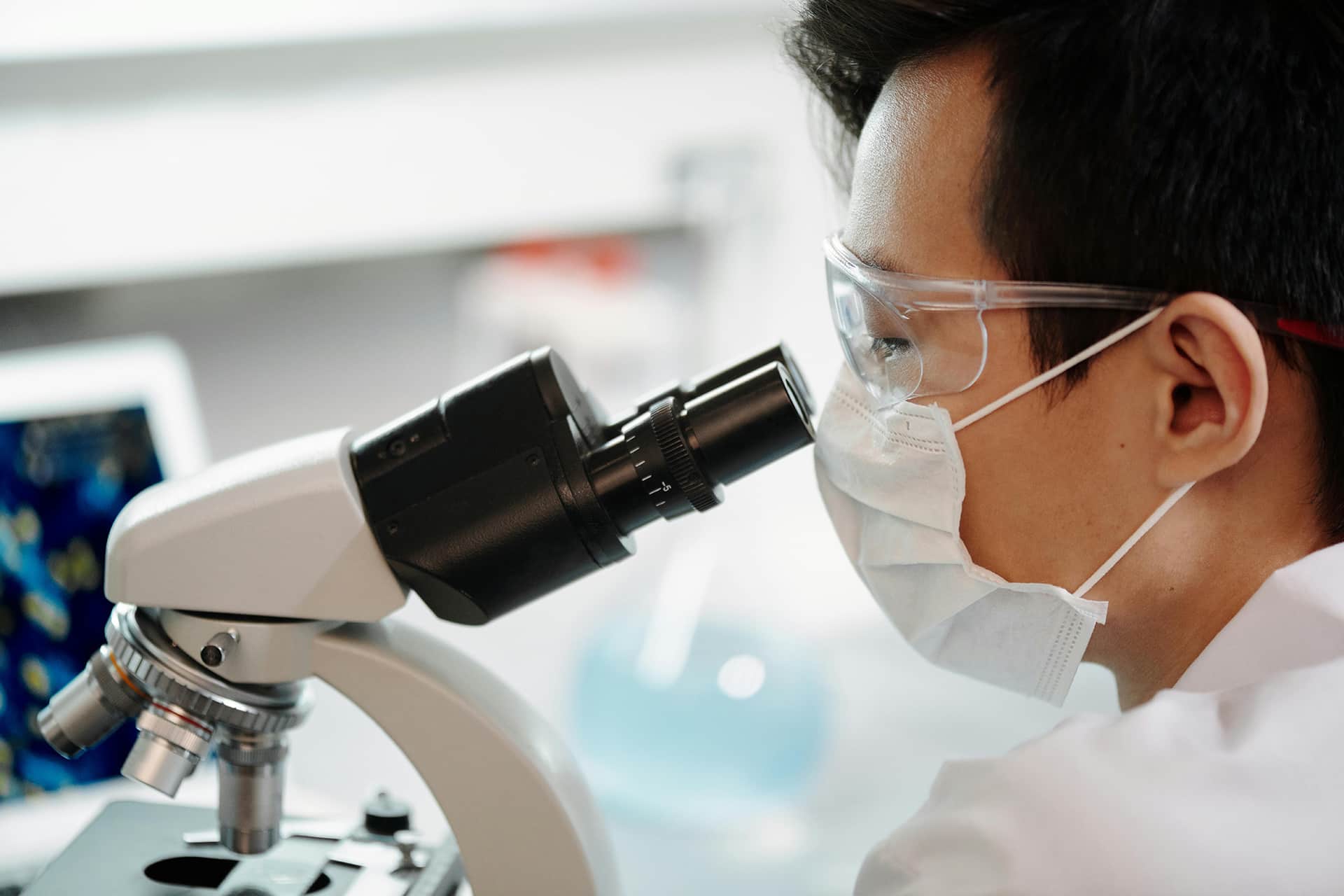 Scientist wearing protective glasses and a face mask while observing through a microscope in a laboratory